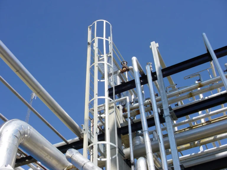 Galvanized caged ladder rising through process piping against a blue sky