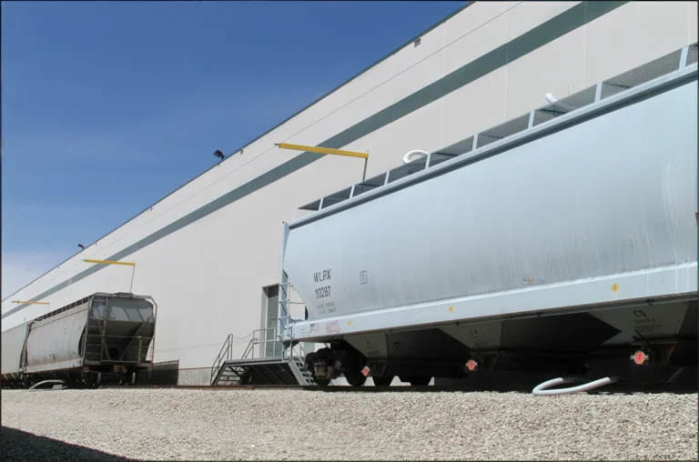 Hopper railcars along a warehouse wall with wall-mounted yellow fall arrest arms and access stairs
