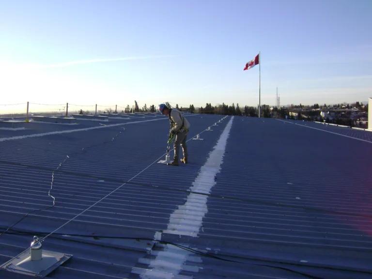 Worker tied off to a horizontal lifeline on a large low-slope metal roof at dusk